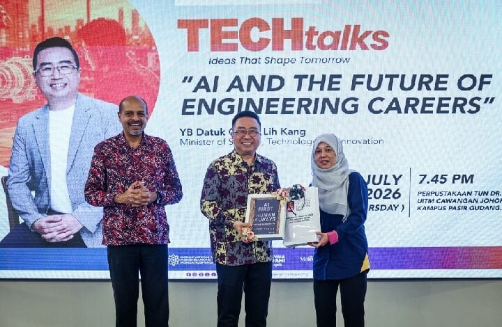 mosti 2 - Wilayah.com.my Three people on a stage during a TEC talks event; a man in patterned shirt receives a book and a bag from a woman in a hijab, with a large screen in the background showing the event title.