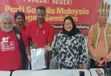 muda - Wilayah.com.my Group of five at a campaign event; a man in red hands a document to a smiling woman in a black hijab, with a banner behind them.