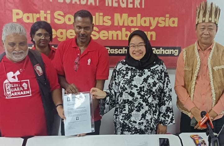 muda - Wilayah.com.my Group of five at a campaign event; a man in red hands a document to a smiling woman in a black hijab, with a banner behind them.