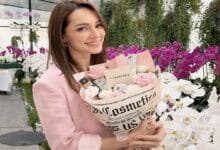 nadia anuar - Wilayah.com.my Smiling woman in a pink blazer holds a bouquet of pink and white roses wrapped in newspaper print, in a bright flower shop.