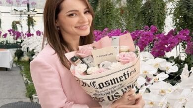 nadia anuar - Wilayah.com.my Smiling woman in a pink blazer holds a bouquet of pink and white roses wrapped in newspaper print, in a bright flower shop.