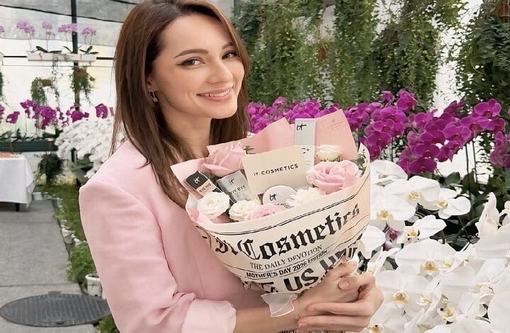 nadia anuar - Wilayah.com.my Smiling woman in a pink blazer holds a bouquet of pink and white roses wrapped in newspaper print, in a bright flower shop.