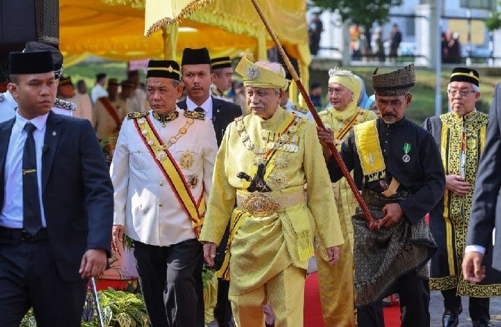 negeri sembilan - Wilayah.com.my Group of dignitaries in ornate ceremonial outfits walking on a red carpet; central figure wears gold attire and a crown.