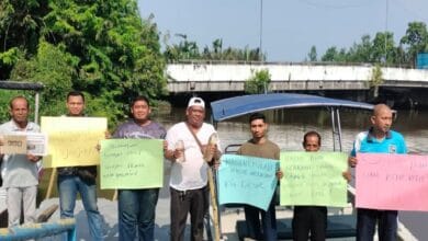 nelayan - Wilayah.com.my Eight men stand on a riverside walkway holding colorful handwritten protest signs, with a blue boat and a bridge in the background.