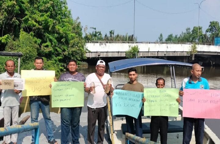 nelayan - Wilayah.com.my Eight men stand on a riverside walkway holding colorful handwritten protest signs, with a blue boat and a bridge in the background.