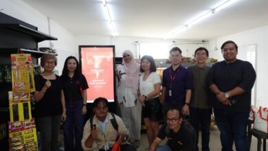 ninja mart - Wilayah.com.my Group of nine adults posing for a photo in a bright room, some with ID badges, a red display screen behind them suggesting a training or event origin.
