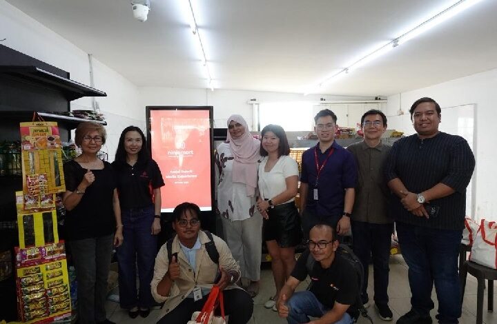 Group of nine adults posing for a photo in a bright room, some with ID badges, a red display screen behind them suggesting a training or event origin.