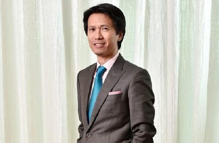 Professional portrait of a man in a gray suit, teal tie, and pink pocket square posing with hands in pockets against a light curtain backdrop.