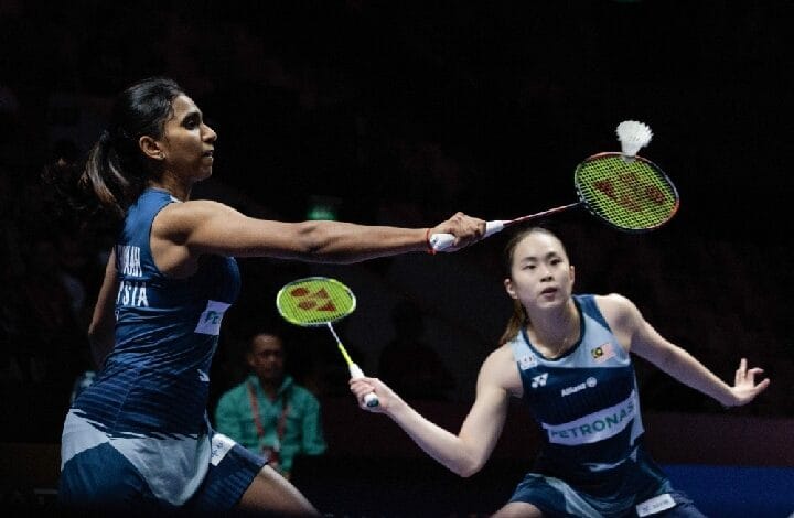 Two female badminton players in blue uniforms stretch to return the shuttle during a doubles match.