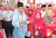 ph - Wilayah.com.my Man in a light blue traditional outfit and black cap speaks to a crowd at a red campaign rally, surrounded by supporters in red with lanyards.