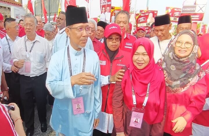 ph - Wilayah.com.my Man in a light blue traditional outfit and black cap speaks to a crowd at a red campaign rally, surrounded by supporters in red with lanyards.