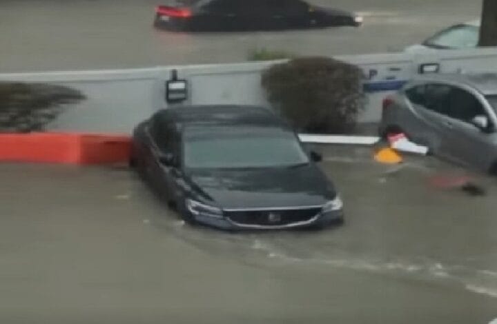 pj banjir 2 - Wilayah.com.my Dark sedan partially submerged in floodwater on a street, with another car nearby and a police vehicle in the background.