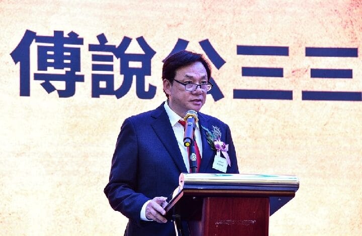 poh po lian - Wilayah.com.my Man in a dark suit and glasses giving a speech at a podium with a microphone, flower boutonniere, in front of a large Chinese character backdrop.