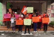 psm - Wilayah.com.my Group of diverse protesters on a storefront porch, holding colorful signs and a red flag, raising fists in a demonstration.