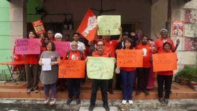 psm - Wilayah.com.my Group of diverse protesters on a storefront porch, holding colorful signs and a red flag, raising fists in a demonstration.