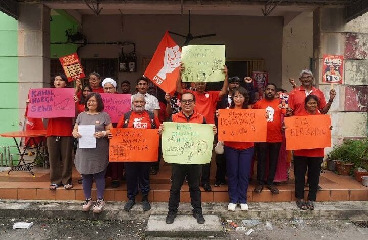 psm - Wilayah.com.my Group of diverse protesters on a storefront porch, holding colorful signs and a red flag, raising fists in a demonstration.