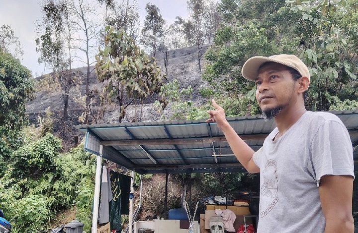 Man wearing a beige cap points toward a burnt hillside behind a small shelter and trees in an outdoor setting while looking to the side.