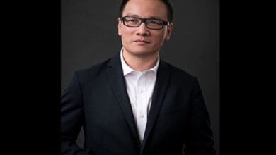 Professional headshot of an Asian man in a dark suit and white shirt, wearing glasses, against a dark background.