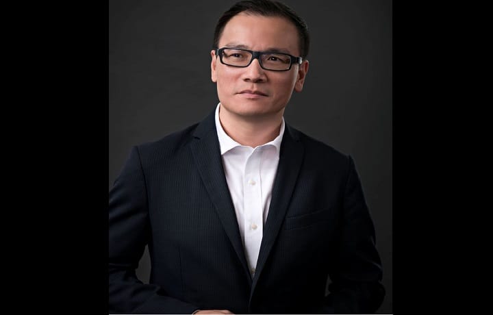 raymond tan - Wilayah.com.my Professional headshot of an Asian man in a dark suit and white shirt, wearing glasses, against a dark background.