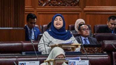 risda - Wilayah.com.my Woman wearing a blue hijab speaking at a lectern in a wood-paneled senate chamber, surrounded by seated colleagues.