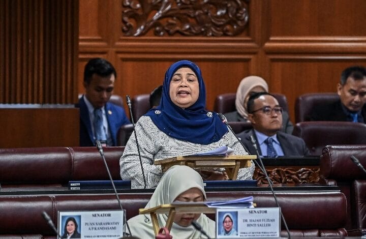 Woman wearing a blue hijab speaking at a lectern in a wood-paneled senate chamber, surrounded by seated colleagues.