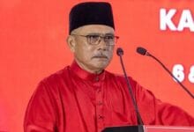 ronald kiandee - Wilayah.com.my Older man in a red traditional outfit and black cap delivering a speech at a podium against a red backdrop