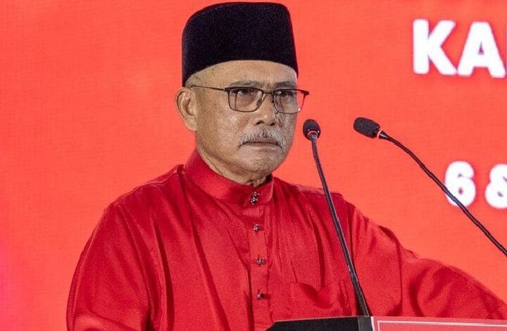 ronald kiandee - Wilayah.com.my Older man in a red traditional outfit and black cap delivering a speech at a podium against a red backdrop