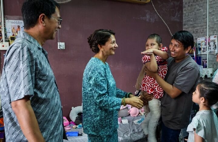 sarafudin badlishah - Wilayah.com.my Woman in teal outfit shakes hands with a man holding his child, as a girl watches in a casual family room setting.