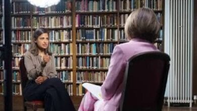 sarah karim khan - Wilayah.com.my Two women sit in a library for an interview: a woman facing the camera speaks with a thoughtful gesture, while another woman with a pink blazer is seen from the back, seated opposite amid tall book shelves.