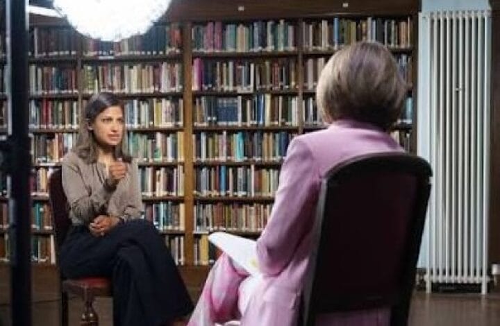 sarah karim khan - Wilayah.com.my Two women sit in a library for an interview: a woman facing the camera speaks with a thoughtful gesture, while another woman with a pink blazer is seen from the back, seated opposite amid tall book shelves.