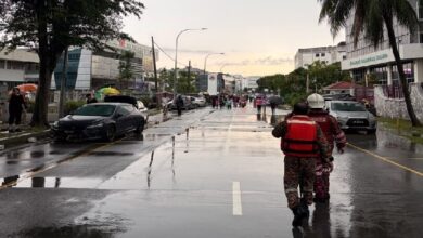selangor banjir kilat - Wilayah.com.my Rainy city street with puddles, pedestrians in the distance, and two responders in red life jackets walking away from the camera; parked cars and buildings line the road.