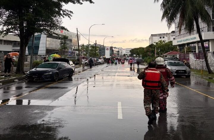 selangor banjir kilat - Wilayah.com.my Rainy city street with puddles, pedestrians in the distance, and two responders in red life jackets walking away from the camera; parked cars and buildings line the road.
