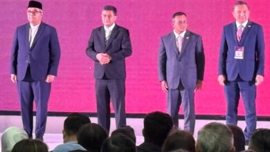 selangor invest 2 - Wilayah.com.my Four men in suits stand on a stage with a bright pink backdrop, facing an audience. The man on the far left wears a cap; the others have hands at their sides or clasped. A row of heads从 the audience is visible in the foreground.