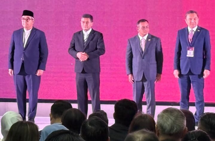 selangor invest 2 - Wilayah.com.my Four men in suits stand on a stage with a bright pink backdrop, facing an audience. The man on the far left wears a cap; the others have hands at their sides or clasped. A row of heads从 the audience is visible in the foreground.