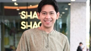 shafiq erwin - Wilayah.com.my Smiling man in a beige kurta with a pendant stands indoors in front of a glass storefront; blurred background shows the letters 'SHA' on a sign.