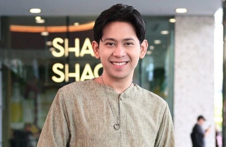 shafiq erwin - Wilayah.com.my Smiling man in a beige kurta with a pendant stands indoors in front of a glass storefront; blurred background shows the letters 'SHA' on a sign.