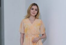 Woman in a peach and yellow pastel wrap dress with a smocked waist, standing against a pale wall indoors.