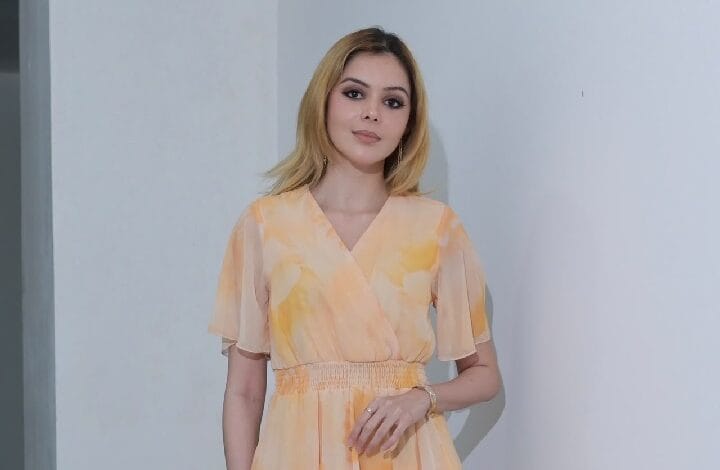 Woman in a peach and yellow pastel wrap dress with a smocked waist, standing against a pale wall indoors.