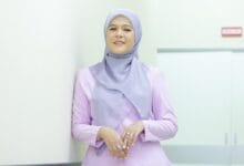 Woman wearing a light purple hijab and lavender top, standing against a white wall in a bright corridor, smiling at the camera.