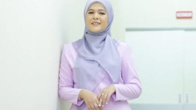 Woman wearing a light purple hijab and lavender top, standing against a white wall in a bright corridor, smiling at the camera.