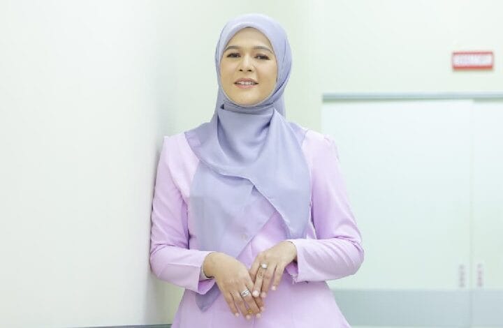 sharifah zarina - Wilayah.com.my Woman wearing a light purple hijab and lavender top, standing against a white wall in a bright corridor, smiling at the camera.