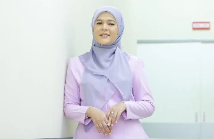 sharifah zarina - Wilayah.com.my Woman wearing a light purple hijab and lavender top, standing against a white wall in a bright corridor, smiling at the camera.