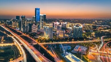 Aerial cityscape at twilight with tall glass skyscrapers and a glowing network of highways and streets.