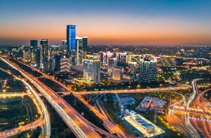 Aerial cityscape at twilight with tall glass skyscrapers and a glowing network of highways and streets.
