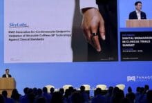 Speaker at a podium on a blue conference stage, a large slide reads about wearable cuffless BP technology for cardiovascular endpoints; audience seated in front.