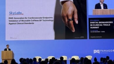 Speaker at a podium on a blue conference stage, a large slide reads about wearable cuffless BP technology for cardiovascular endpoints; audience seated in front.