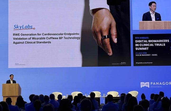sky labs 2 - Wilayah.com.my Speaker at a podium on a blue conference stage, a large slide reads about wearable cuffless BP technology for cardiovascular endpoints; audience seated in front.