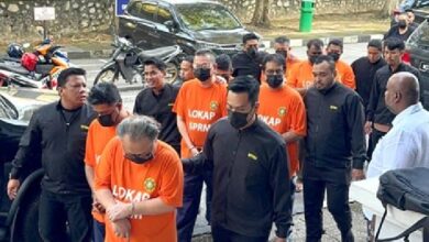 sprm - Wilayah.com.my Group of men in orange detainee shirts labeled LOKAP SPRM, escorted by officials in black outfits; outdoors near parked cars and motorcycles.