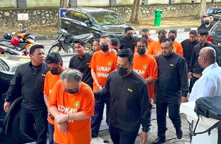 sprm - Wilayah.com.my Group of men in orange detainee shirts labeled LOKAP SPRM, escorted by officials in black outfits; outdoors near parked cars and motorcycles.