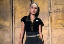stacy 2 - Wilayah.com.my Woman in a black outfit with gold jewelry standing against a rustic wooden door backdrop.